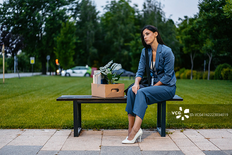 伤心的年轻女商人坐在公司大楼前的长椅上，手里拿着一箱办公用品。图片素材