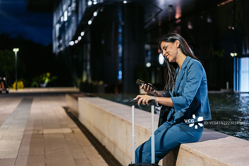 年轻女商人在夜晚坐在公司大楼前使用手机，身旁有行李箱。图片素材