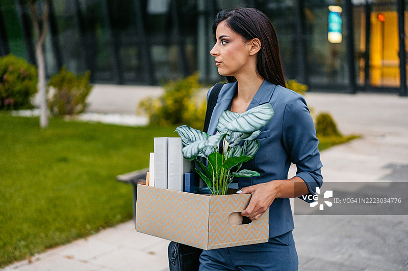 伤心的年轻女商人手拿办公桌物品的盒子，走出一座公司大楼图片素材