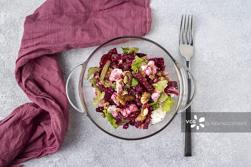 玻璃盘中的甜菜沙拉、绿叶、羊奶酪和核桃，旁边有一条酒红色的餐巾和一把叉子，放在灰色的桌子上。图片素材