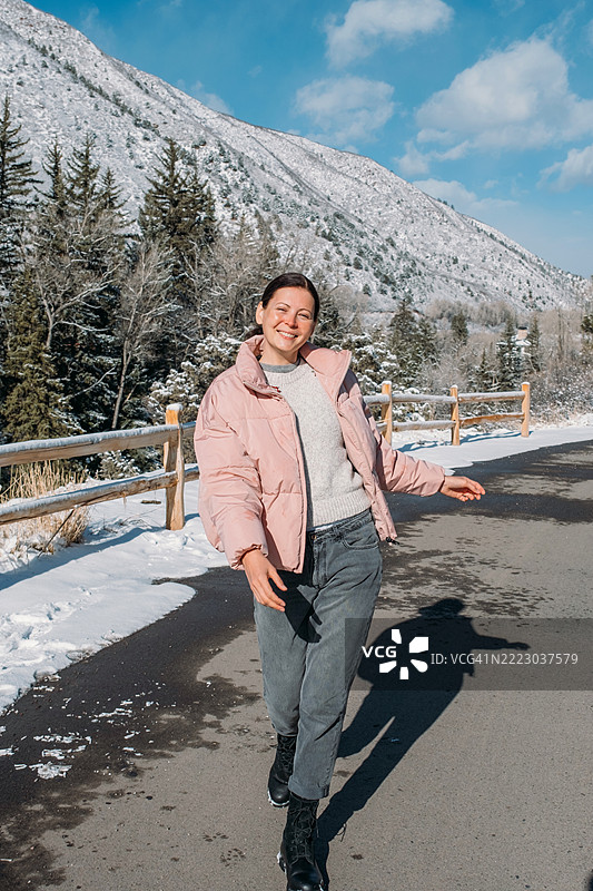 快乐的女人在雪山旁的清晰道路上行走。阳光明媚，地面上有雪，背景中有树木。冬季旅行与幸福的概念。图片素材