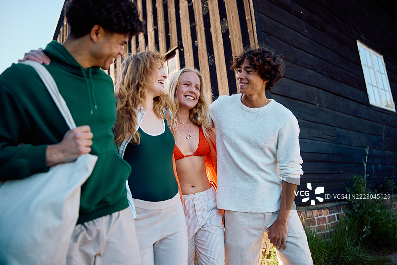 一群年轻朋友穿着夏季服装，微笑着沿着小路走向一座乡村木屋。图片素材