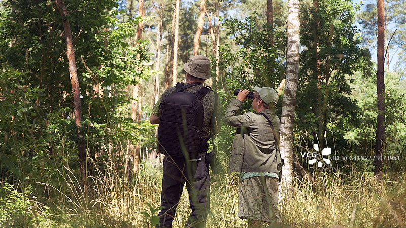 父子在绿色的夏季森林中，周围是树木和高草。鸟类观察家庭冒险。通过望远镜观察鸟类。图片素材