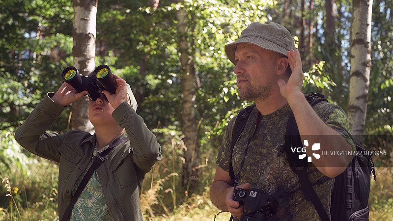父子在绿树成荫的森林中，享受鸟类观察的家庭冒险。他们正在通过望远镜观察鸟类。图片素材