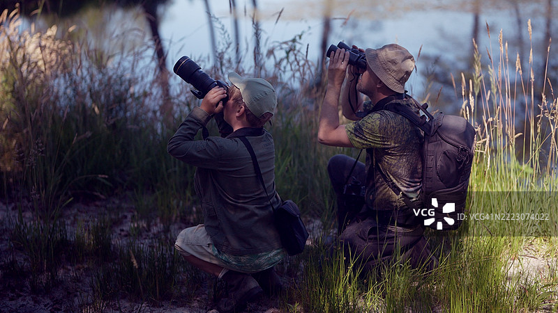 父亲和儿子在郁郁葱葱的夏季森林中，清澈蓝湖旁共度时光。家庭鸟类观察冒险，使用望远镜观察并拍照。鸟类观察。图片素材