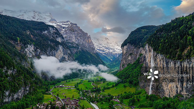 瑞士阿尔卑斯山劳特布伦嫩谷的鸟瞰图,斯陶巴赫瀑布从陡峭的悬崖上倾泻而下,传统的瑞士小木屋坐落在郁郁葱葱的草地上,背景是被雪覆盖的山峰,适合旅游摄影的自然与旅行概念。图片素材