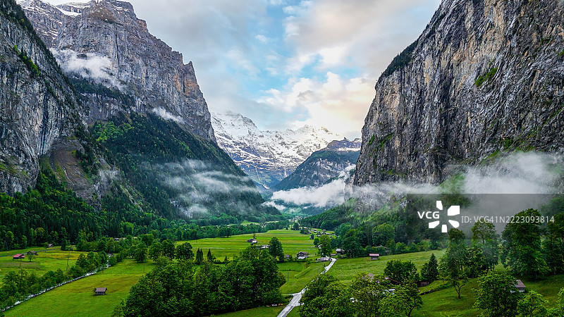 瑞士阿尔卑斯山劳特布伦嫩谷的鸟瞰图,斯陶巴赫瀑布从陡峭的悬崖上倾泻而下,传统的瑞士小木屋坐落在郁郁葱葱的草地上,背景是被雪覆盖的山峰,适合旅游摄影的自然与旅行概念。图片素材