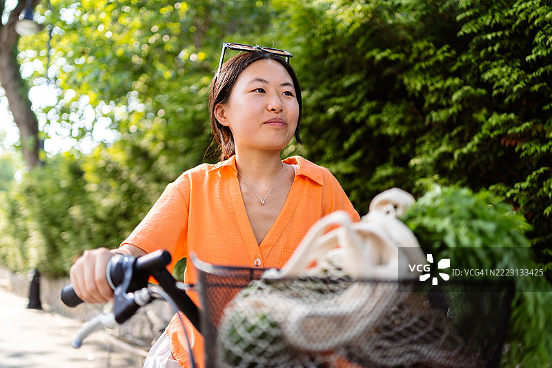 微笑的女性骑着自行车沿着乡村道路行驶，前篮里装着新鲜的杂货，周围是绿色的风景。图片素材