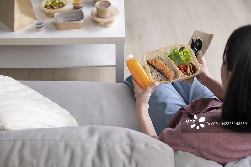年轻的亚洲女性坐在沙发上，在家享用健康餐。图片素材