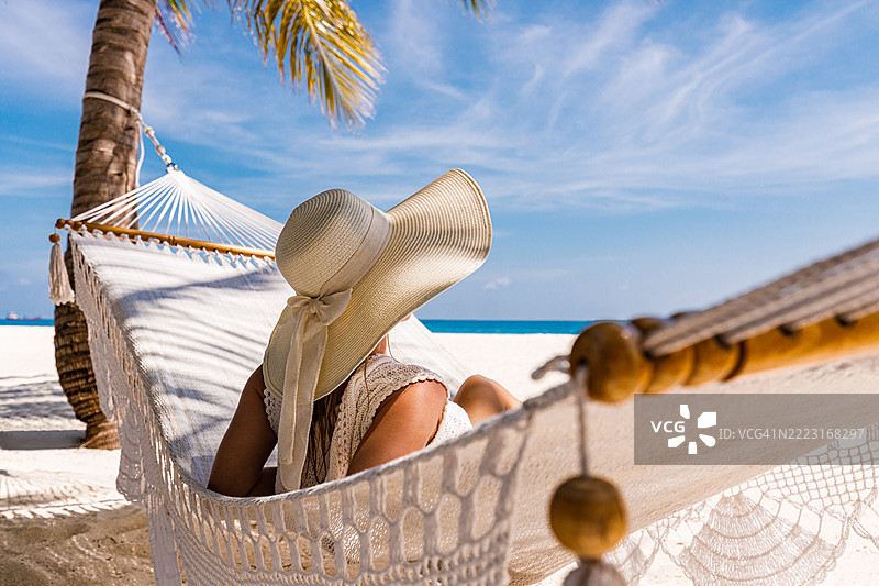 在海滩上，戴着帽子的放松女性躺在吊床上。图片素材
