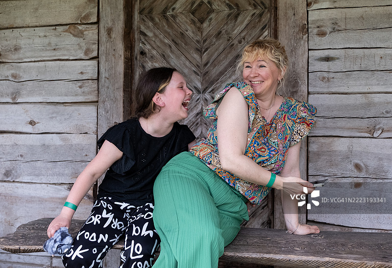一个9岁的白人女孩和一位年轻女性坐在一个古老的木质露台上，笑着聊天。她们手里拿着一部手机。图片素材