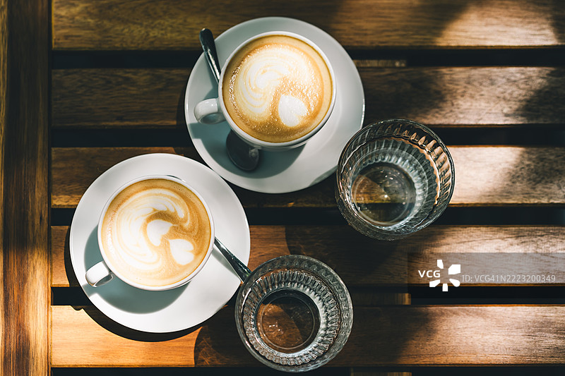 两杯心形拉花的卡布奇诺咖啡放在木桌上，旁边有水杯。俯视图。图片素材