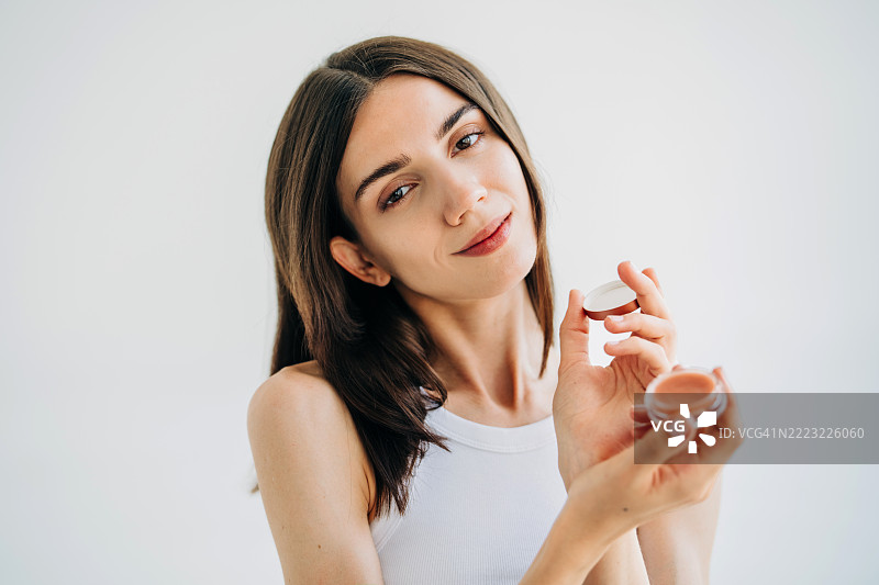 一位美丽女性的特写肖像，温柔地微笑着，手中拿着一个小瓶面霜或润唇膏。美容、护肤和自我护理的概念。图片素材