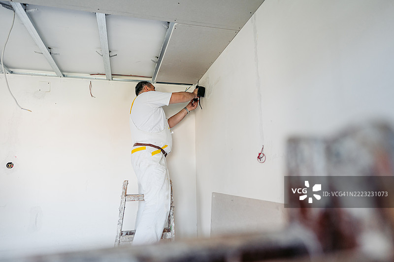 成熟工人在空置的翻新公寓里用电钻安装天花板石膏板图片素材
