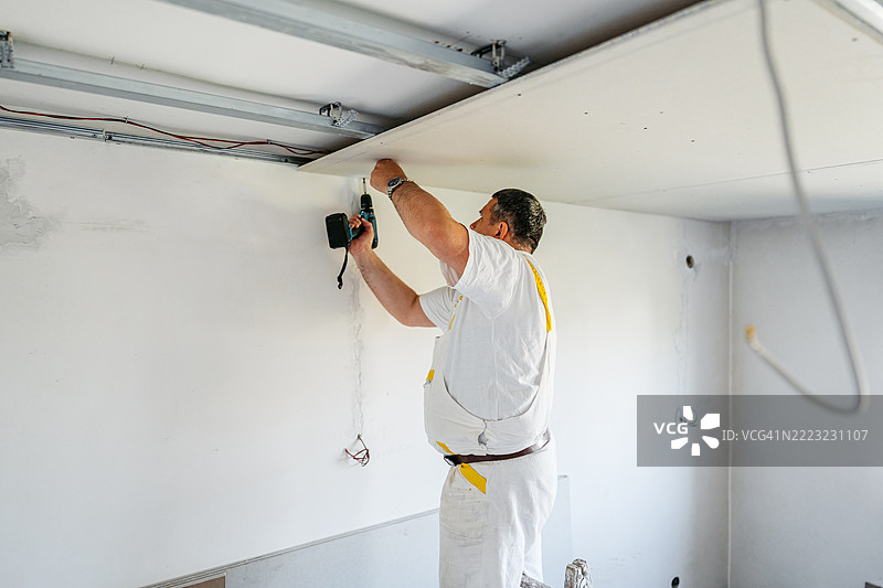 成熟工人在空置的翻新公寓里用电钻安装天花板石膏板图片素材
