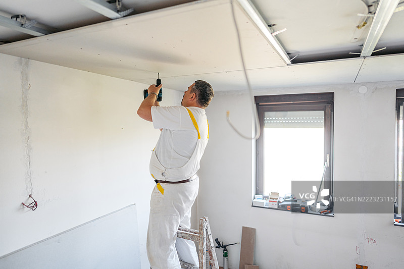 成熟工人在空置的翻新公寓里用电钻安装天花板石膏板图片素材