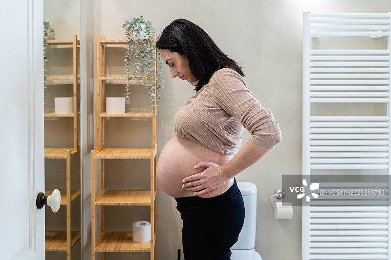 侧面视角的孕妇在浴室里用乳霜滋润她的肚子图片素材