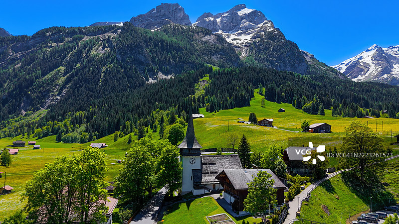 伯尔尼高地的Gsteig bei Gstaad村庄的鸟瞰图,背景是历史悠久的教堂、传统的木屋和白雪覆盖的阿尔卑斯山峰,展现瑞士乡村文化和自然旅游的概念。图片素材