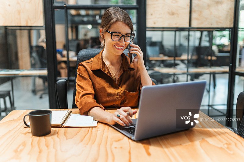 快乐的女商人在共享办公空间的桌子上使用笔记本电脑和手机工作图片素材