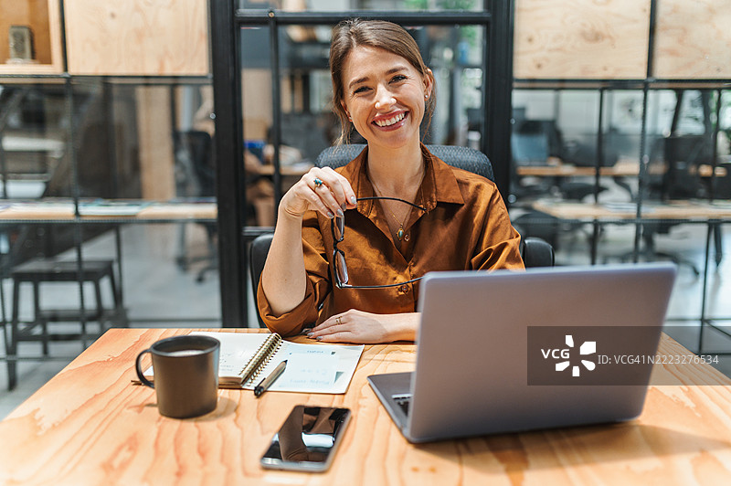微笑的女商人手持眼镜，坐在共享办公空间的工作桌前看向镜头图片素材