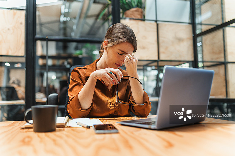 疲惫的女商人在共享办公空间的桌子上，闭着眼睛捏着鼻梁。图片素材