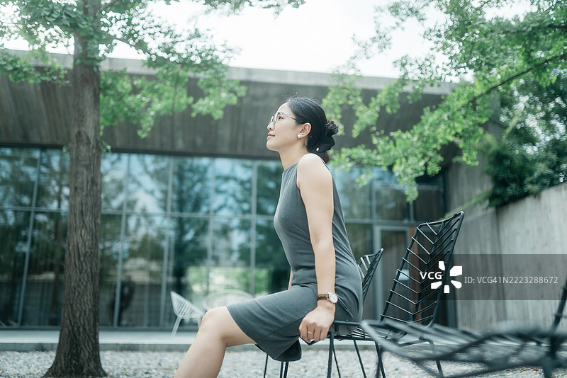 一位迷人的亚洲女性白领穿着商务服装，坐在椅子上，在公园的树下冥想。图片素材