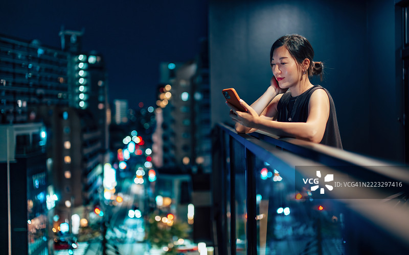 美丽的年轻亚洲女性在公寓阳台上放松，靠在栏杆上滑动智能手机，查看社交媒体。夜晚从高处欣赏城市夜景的繁荣。生活方式与科技图片素材