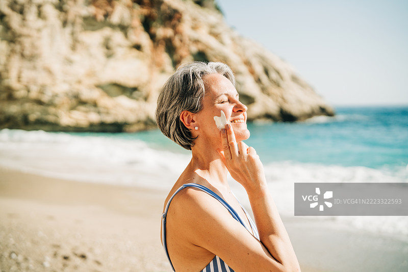 一位快乐的、头发灰白的中年女性在阳光明媚的海滩上，一边笑着，一边在脸颊上涂抹星星形状的防晒霜。图片素材