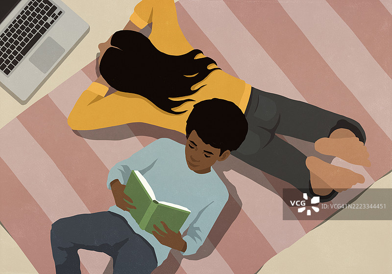 从上方俯视，男孩在家中躺在地板上阅读书籍，母亲则在笔记本电脑上工作。图片素材