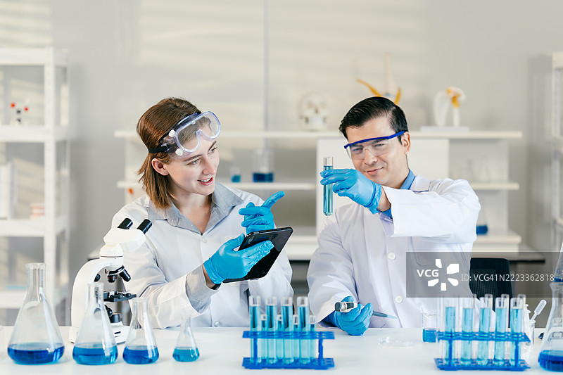 两位穿着安全装备的科学家在现代实验室中合作进行实验，使用平板电脑。研究团队将数字数据分析与实际的化学或生物实验相结合。图片素材