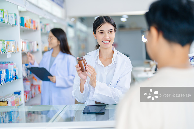 友好的女性药剂师在现代药店中为顾客提供药物建议，背景中还有另一位药剂师。药剂师正在解释产品的好处。图片素材
