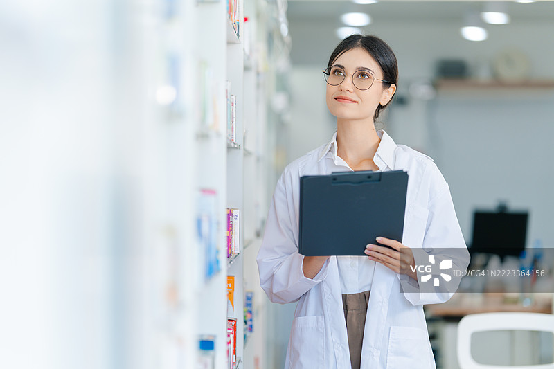 穿着白大褂的女性药剂师在现代药店检查货架上的库存。医疗专业人员正在用夹子整理和核实药店货架上的产品。图片素材