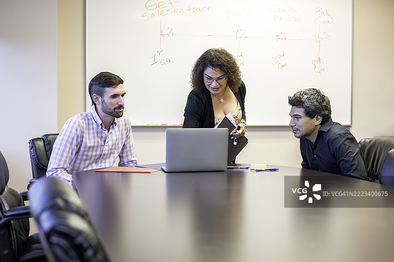 成熟的西班牙裔女性专业办公室工作人员在会议室中使用笔记本电脑为年轻男性同事进行小组演示，指导他们向同事们展示。图片素材