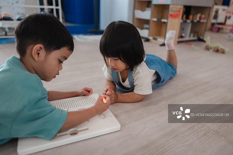两个学龄前的亚洲儿童趴在地板上，一起在白板上画画。图片素材