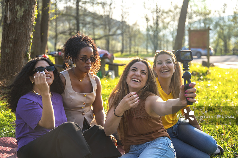 四位不同种族背景的女性朋友坐在公园的草地上，使用稳定器上的运动相机自拍。图片素材