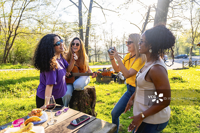 四个开朗的女性朋友正在阳光明媚的公园里野餐，她们弹着吉他、拍照，享受着彼此的陪伴。图片素材