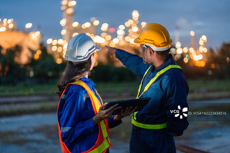 在石油和天然气炼油厂的入口，两名工程师正在使用笔记本电脑。图片素材