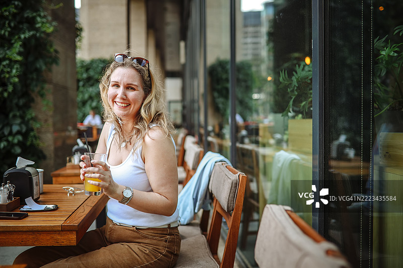 季节：一位快乐的中年女性在街头咖啡馆享用清爽的橙色鸡尾酒，享受城市的夏日温暖。图片素材
