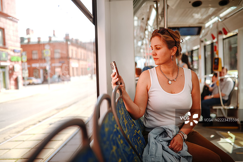美丽的中年女性在城市公共交通上旅行，透过窗户用智能手机欣赏风景。图片素材