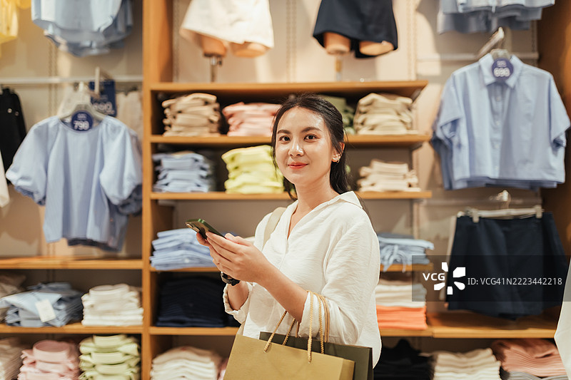亚洲女性站在服装店里，微笑着手持智能手机和购物袋。周围是整齐叠放的衬衫和有序的货架，她享受着购物的乐趣。图片素材
