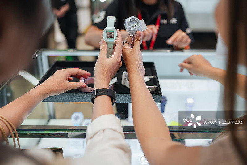 两位女性在现代商店购物智能手表。她们在与销售助理互动的同时比较款式、功能和颜色，展示了生活方式、时尚和消费行为趋势。图片素材