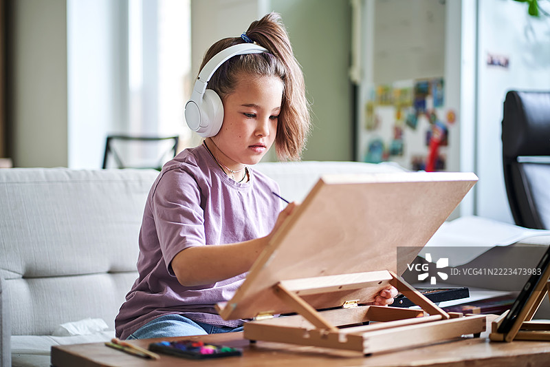 一个戴着耳机的女孩在桌面画架上画画。图片素材