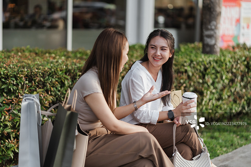 两位亚洲女性在购物后坐在长椅上放松，微笑着聊天，旁边有咖啡和购物袋。这是一个展现友谊、休闲和现代消费文化的自然生活瞬间。图片素材