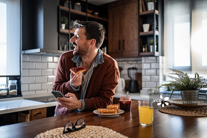 微笑的男人在现代厨房享受早餐和社交媒体图片素材