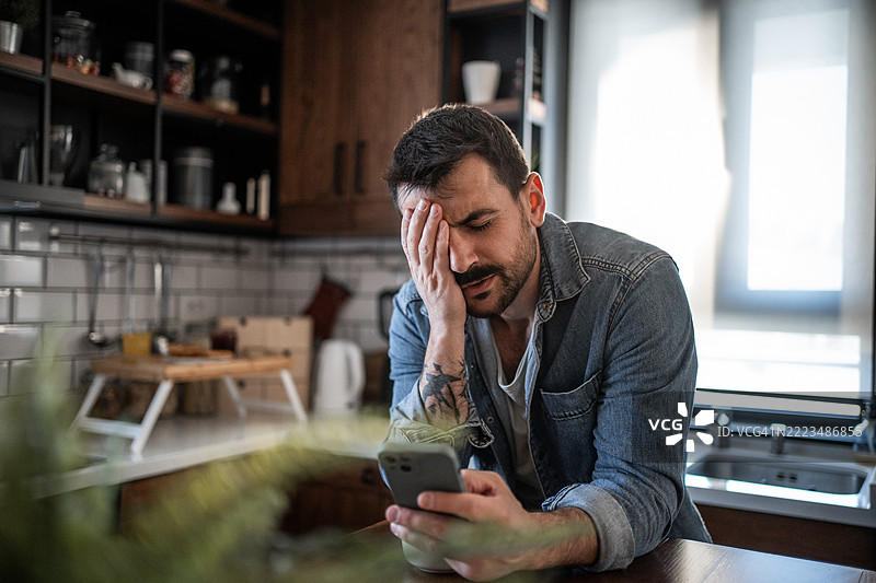 在厨房里，感到压力的男人正在手机上阅读坏消息图片素材
