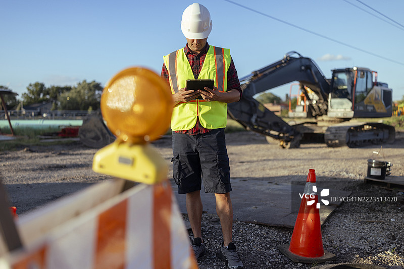 一名工人在检查建筑工地。图中还显示了一个顶部有闪光灯的交通锥或桶，通常用于建筑区域或指挥交通。图片素材