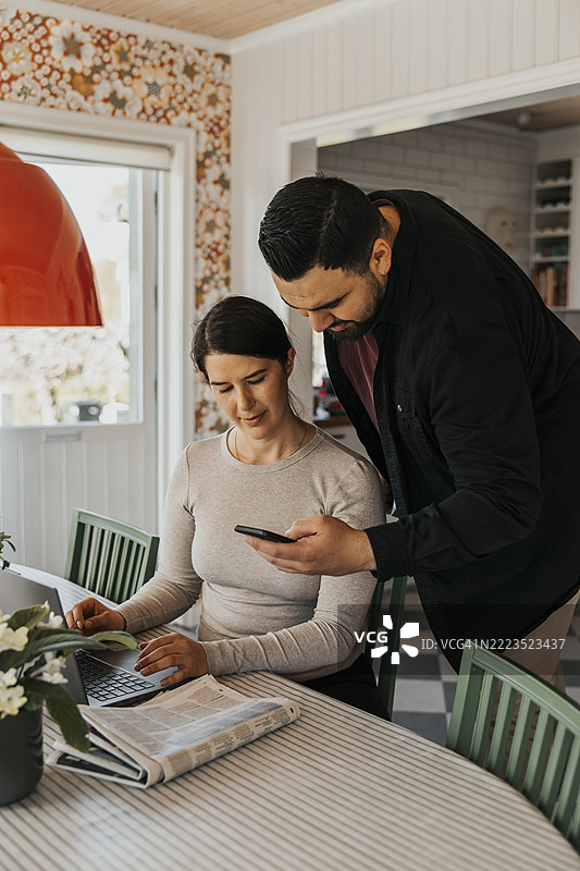 男人在家中餐桌旁向坐着的女人展示智能手机，桌上还有一台笔记本电脑。图片素材