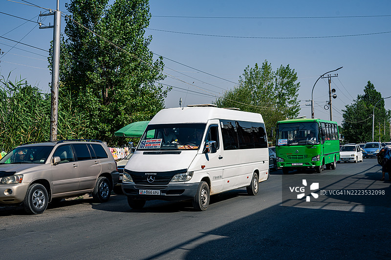 一辆白色梅赛德斯-奔驰斯宾特小巴和一辆小型绿色汽车在吉尔吉斯斯坦奥什的道路上行驶，背景中可见其他车辆和街头小贩。图片素材