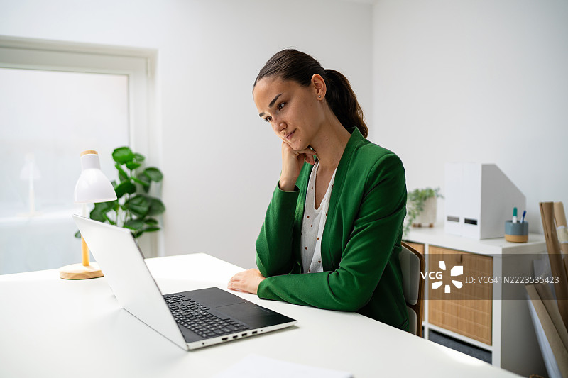 在明亮的办公室空间里，思考的女性坐在桌子前使用笔记本电脑。图片素材