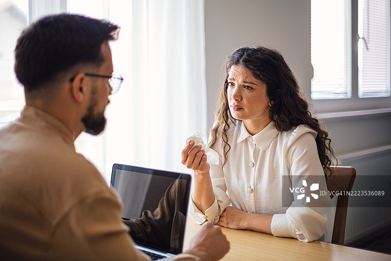 年轻女性拿着纸巾与桌子上的顾问交谈，桌上有一台笔记本电脑。图片素材